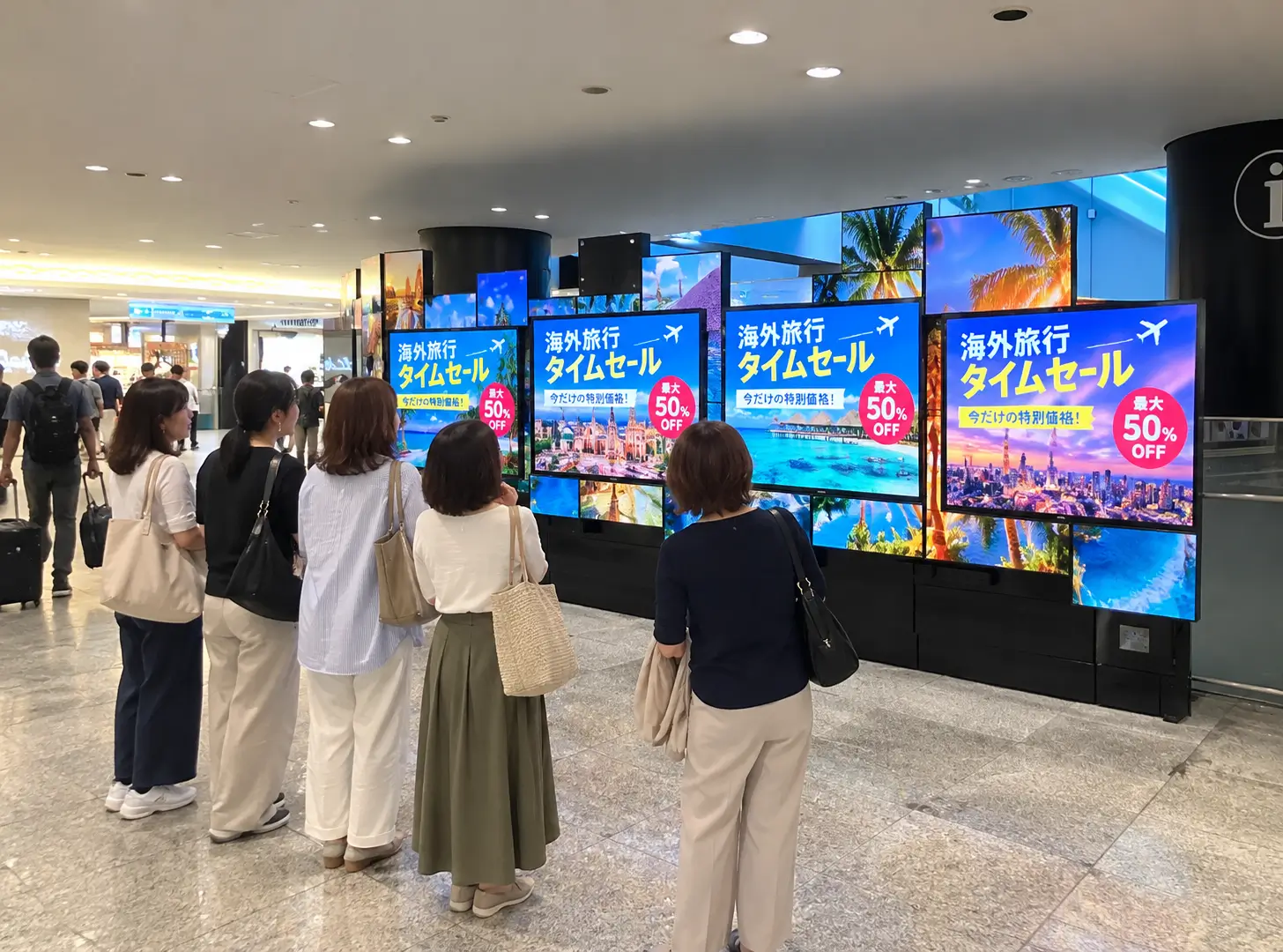 駅構内のマルチ画面デジタルサイネージで流れる海外旅行タイムセール広告に見入る女性グループ