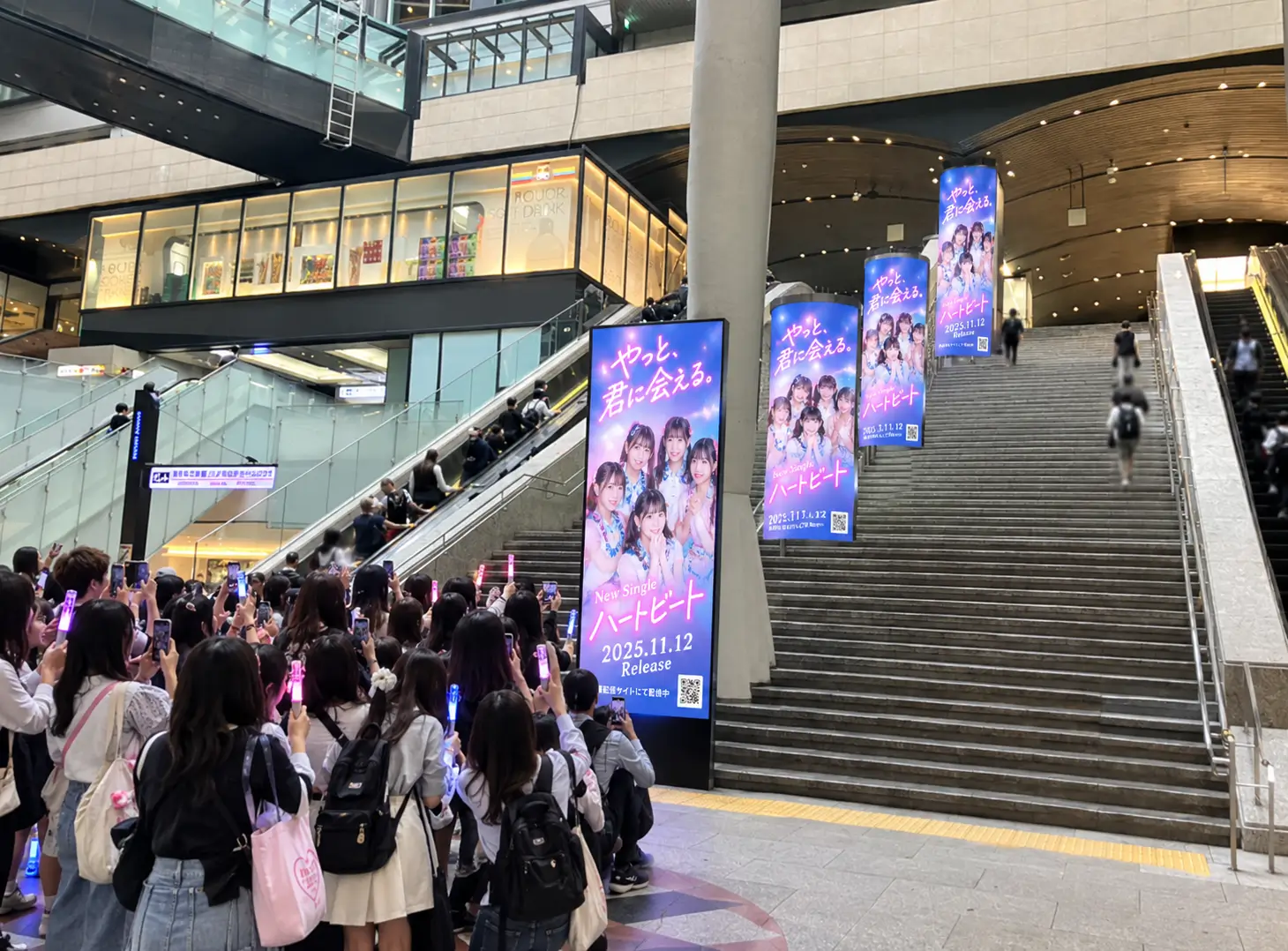 駅の大階段やサイネージに掲出されたアイドルのリリース広告をスマートフォンで撮影するファンたち(SNS拡散・バズの様子)