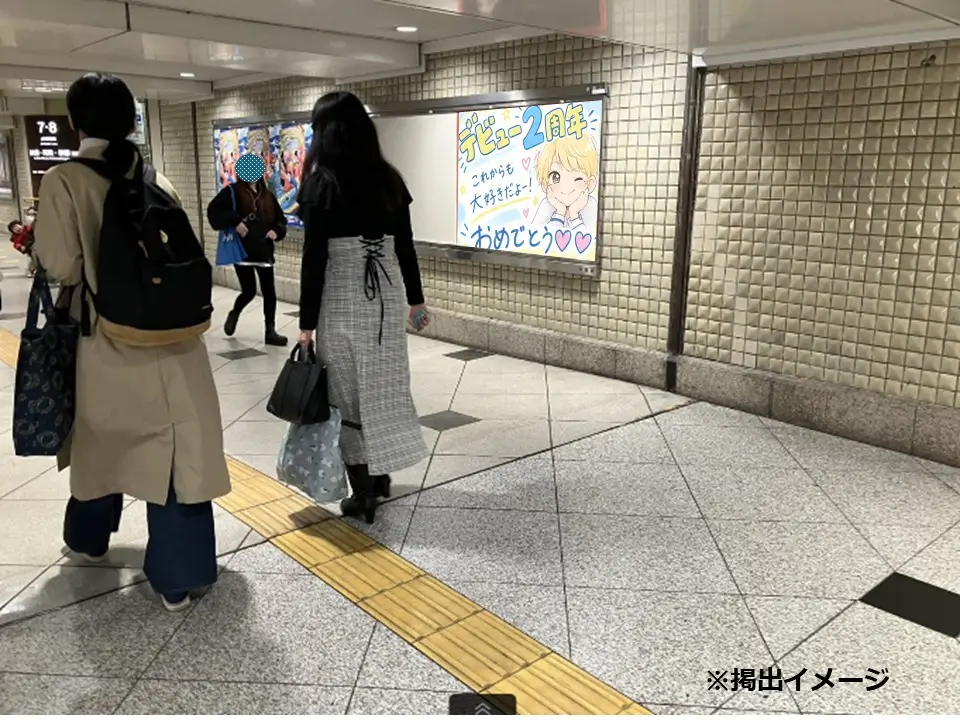 地下鉄通路を行き交う人々と壁面に掲出されたデビュー2周年祝いの応援広告ポスター（遠景）